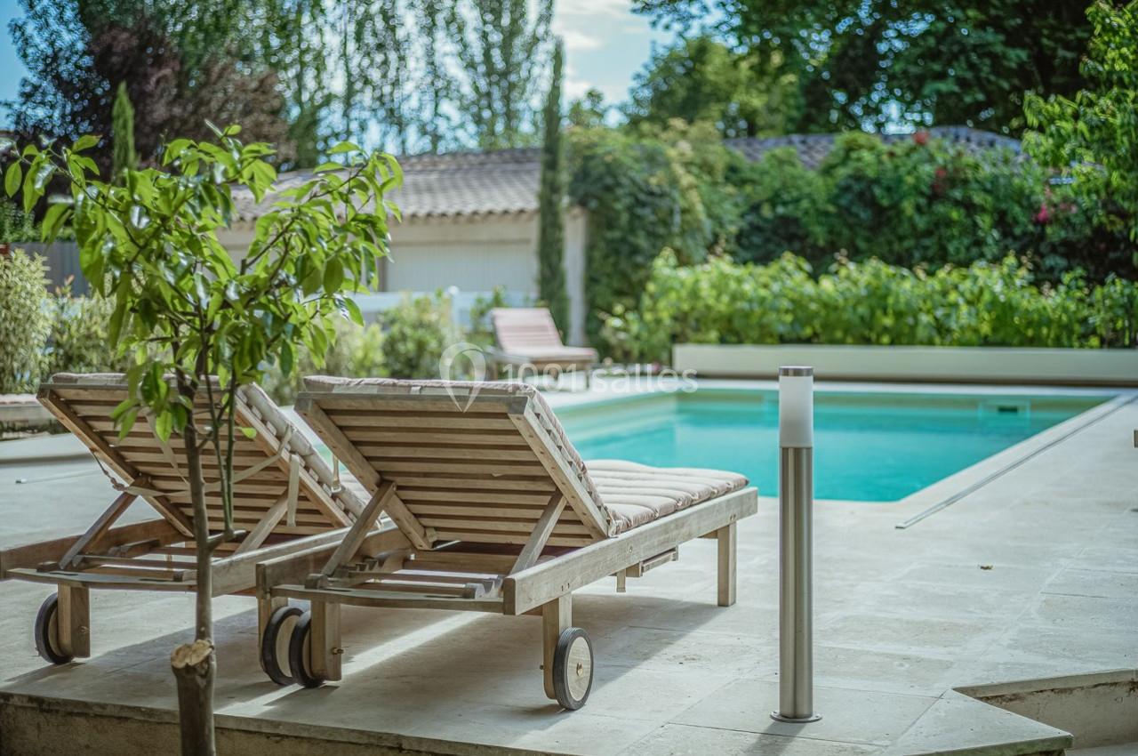 Deux chaises longues en bois avec coussins, placées près d'une piscine entourée de végétation et d'arbres.