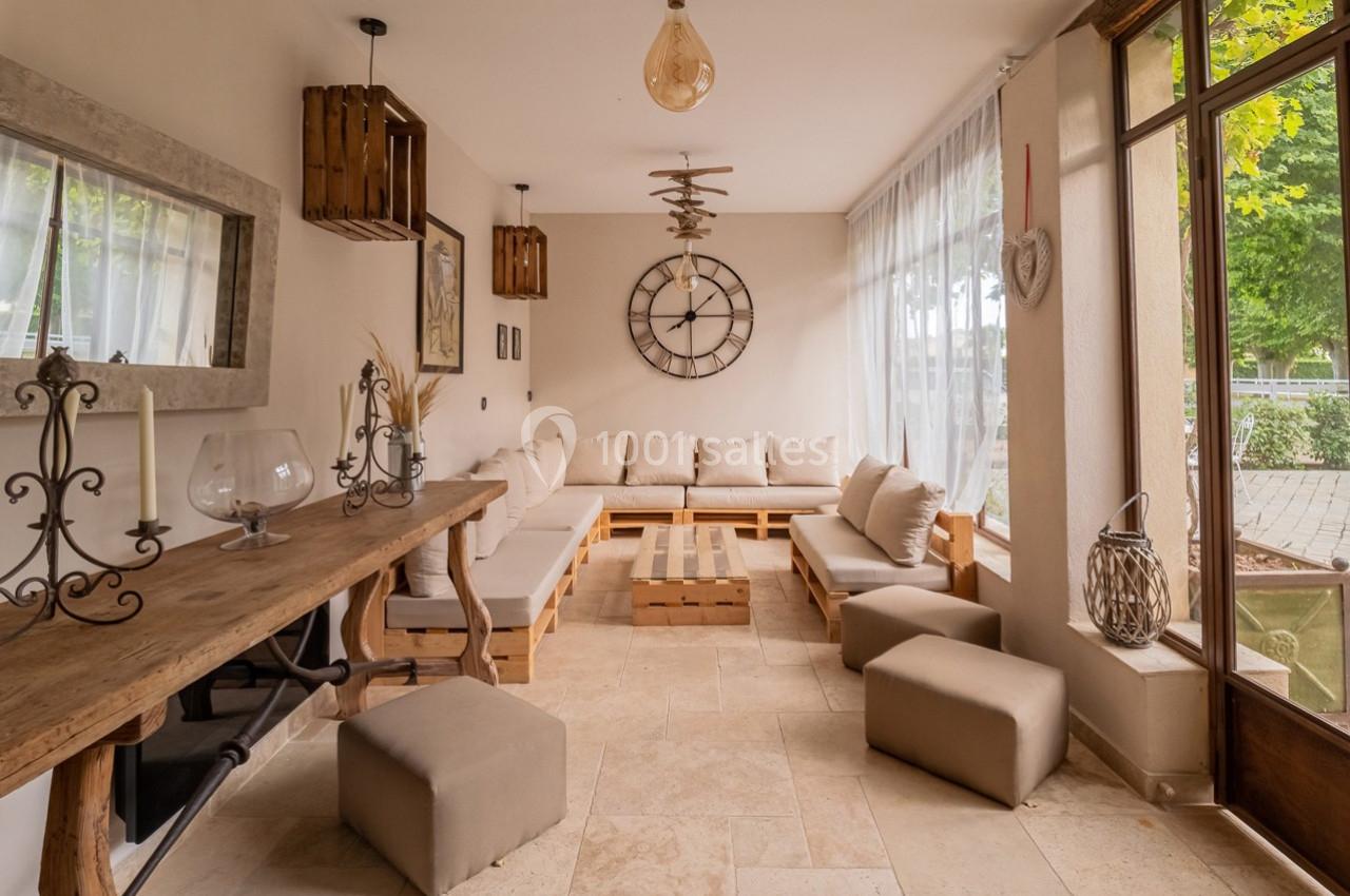 Salon lumineux avec canapé d'angle beige, table basse en bois, décoration murale et grandes fenêtres donnant sur l'extérieur.