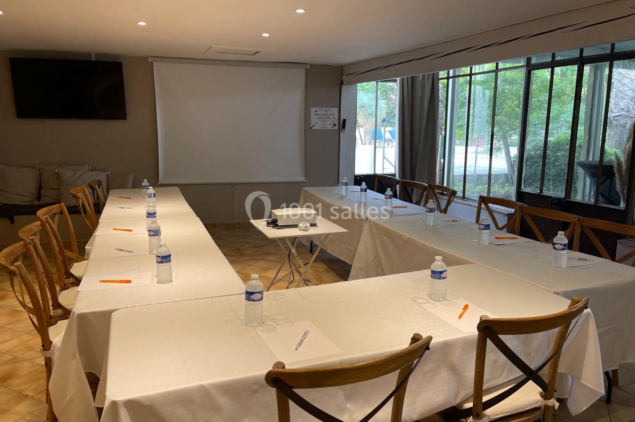 Salle de réunion lumineuse avec tables en U, chaises en bois, bouteilles d'eau, stylos et écran de projection.