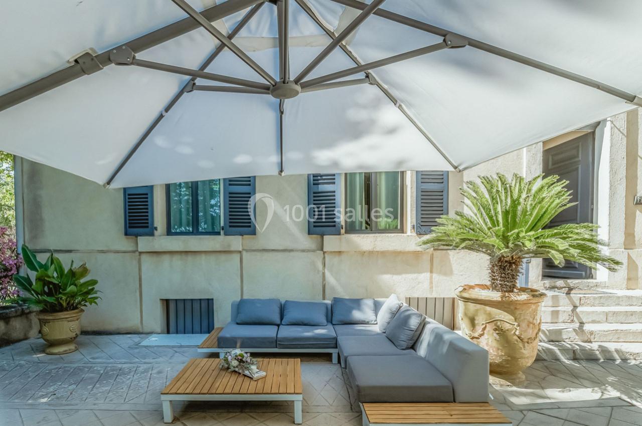 Salon de jardin avec canapé gris, table basse en bois et grand parasol blanc, situé sur une terrasse ensoleillée.