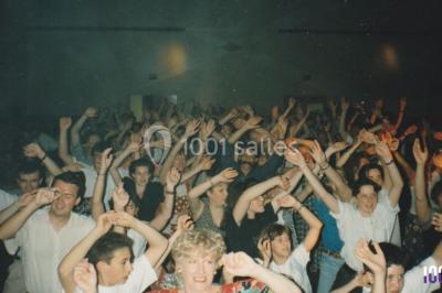 Salle sombre avec des poutres en bois, éclairée par des lumières colorées, des personnes discutent et se déplacent.