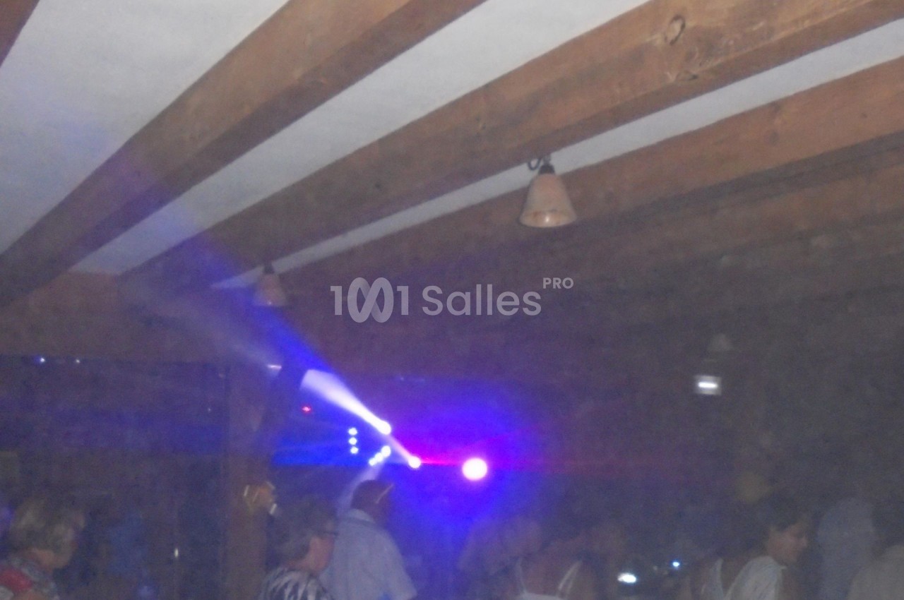 Salle sombre avec des poutres en bois, éclairée par des lumières colorées, des personnes discutent et se déplacent.