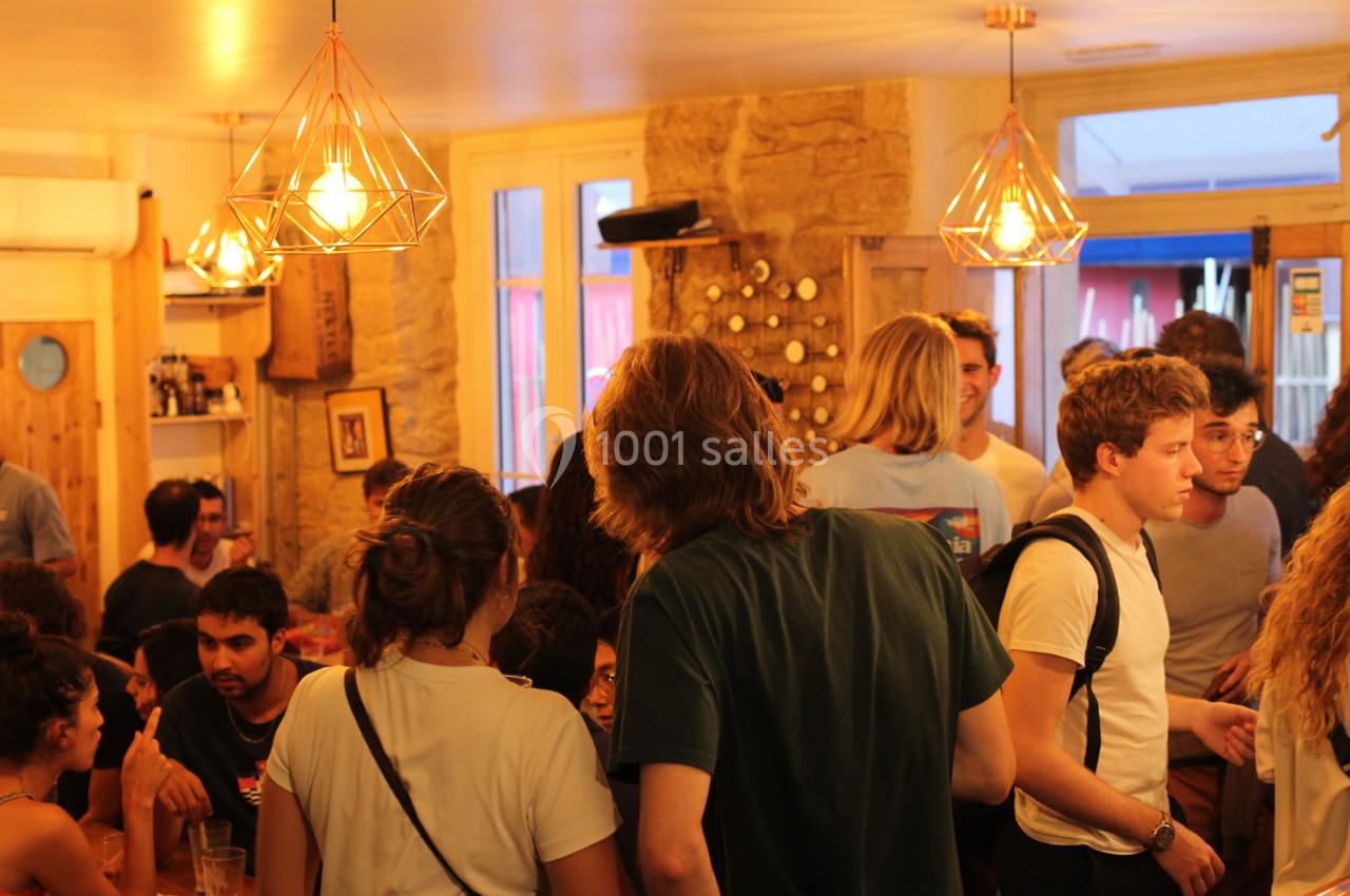 Groupe de personnes discutant dans un café animé, éclairé par des suspensions lumineuses et des murs en pierre.