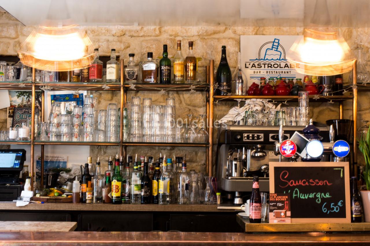 Comptoir de bar avec étagères remplies de bouteilles, verres, machine à café et ardoise indiquant ’Saucisson Auvergne 6,50€’.