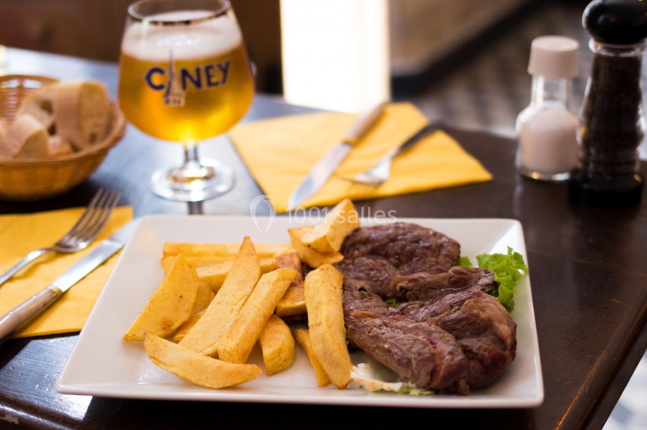 Assiette contenant un steak grillé, des frites et une feuille de salade, accompagnée d'une bière et de pain sur une table.