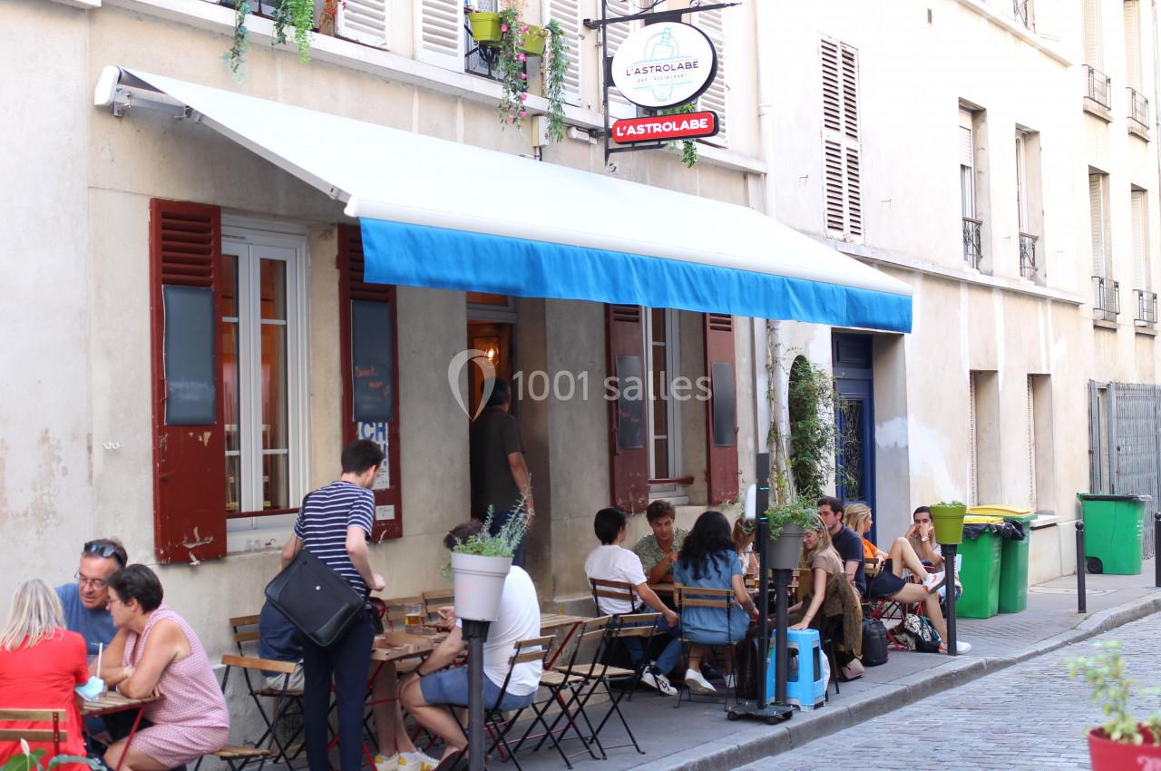 Terrasse d'un café avec des clients assis à des tables, située dans une rue calme bordée d'immeubles.