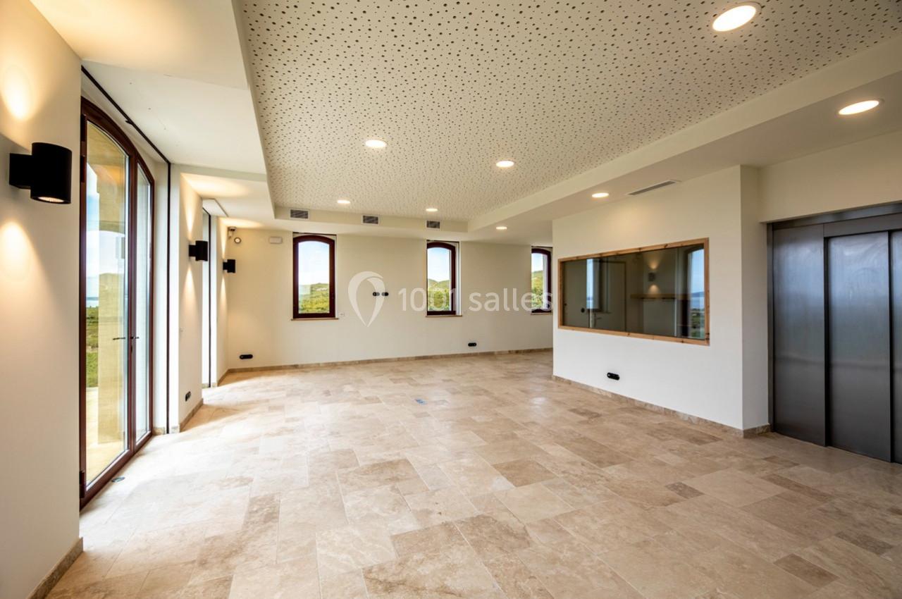 Salle lumineuse avec sol en pierre, grandes fenêtres, plafond perforé, ascenseur et vue sur un paysage extérieur.
