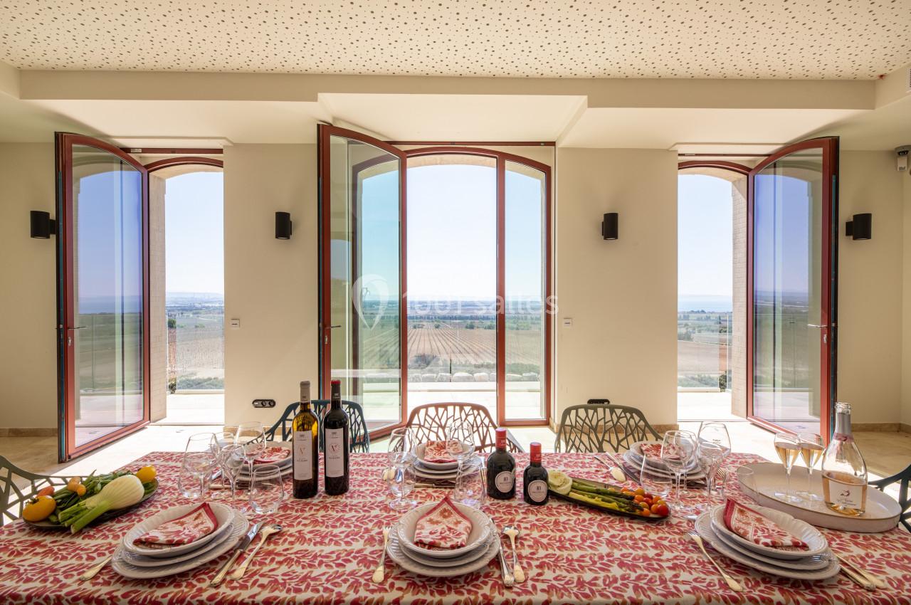 Table dressée avec vaisselle colorée, vue sur un paysage rural à travers de grandes baies vitrées ouvertes.