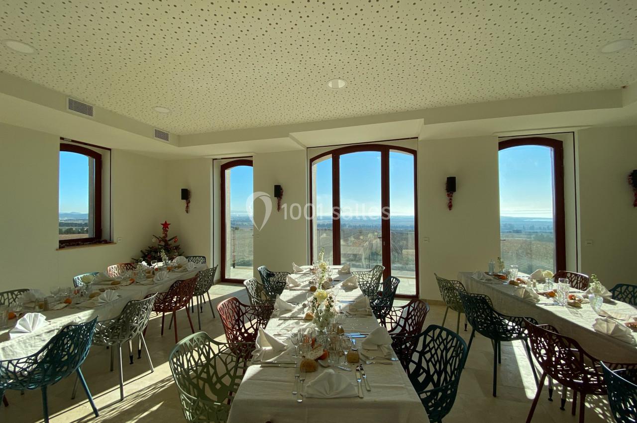 Salle lumineuse avec de grandes fenêtres, tables dressées pour un repas, chaises colorées et vue sur un paysage dégagé.
