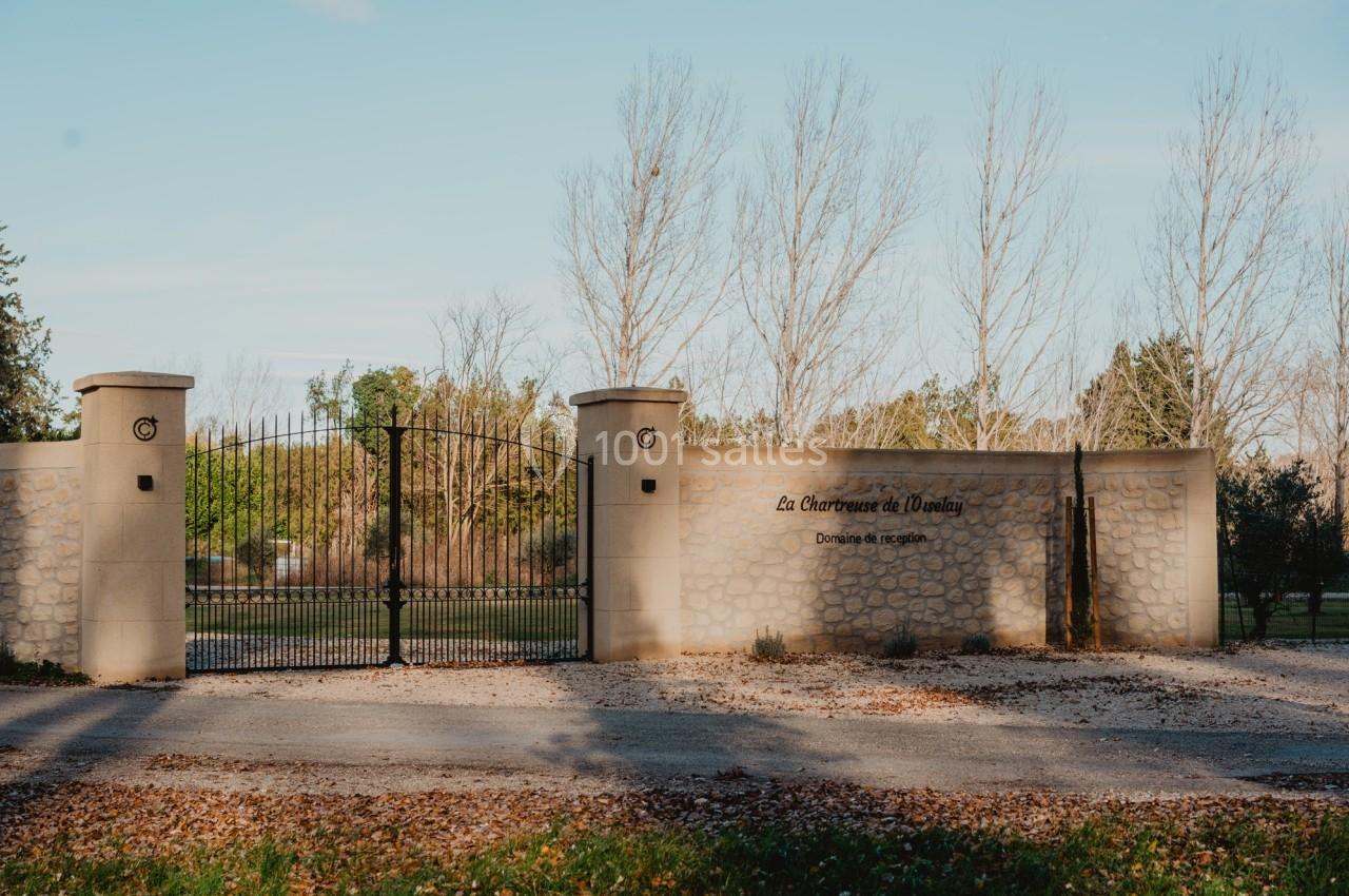 Entrée d'un domaine avec un portail en fer forgé et un mur en pierre, entouré d'arbres et de végétation.