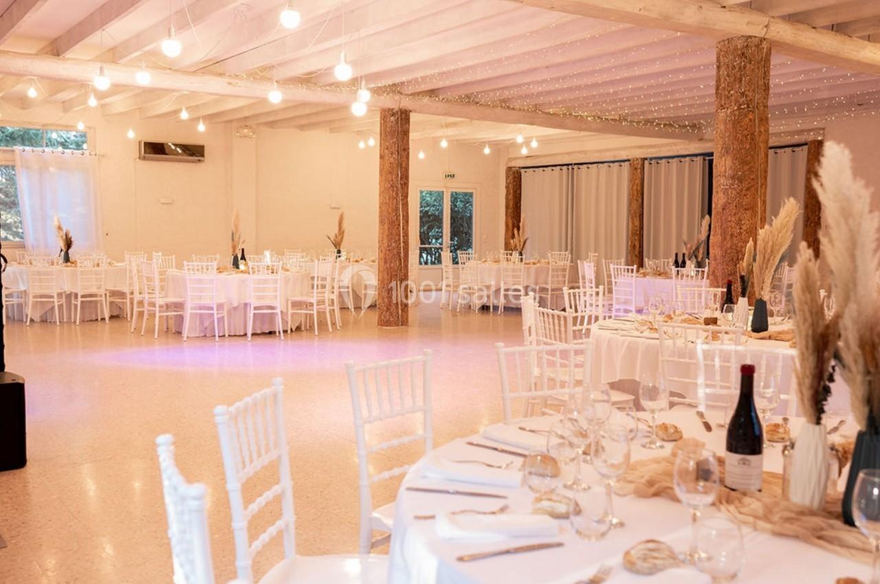 Salle de réception lumineuse avec tables rondes dressées, chaises blanches et décorations naturelles.