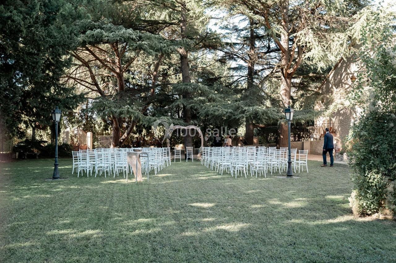 Chaises blanches disposées en rangées sur une pelouse, face à une arche décorative sous de grands arbres.