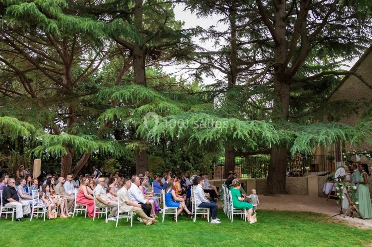Un groupe de personnes assises en extérieur, sous des arbres, assistant à une cérémonie près d'un bâtiment en pierre.