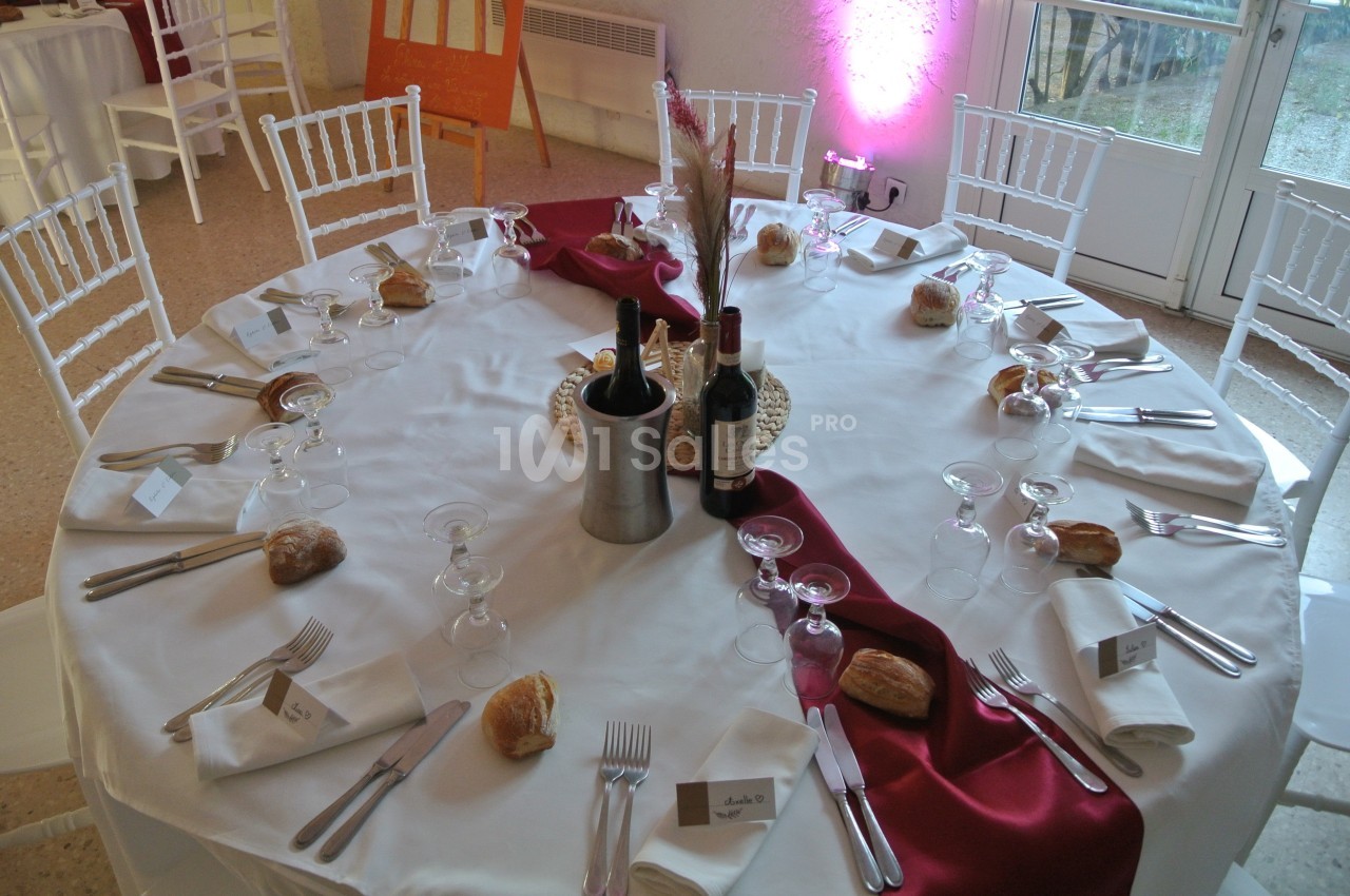 Table ronde dressée pour un repas, avec nappes blanches, chemins de table rouges, couverts, verres, pain et bouteilles de…