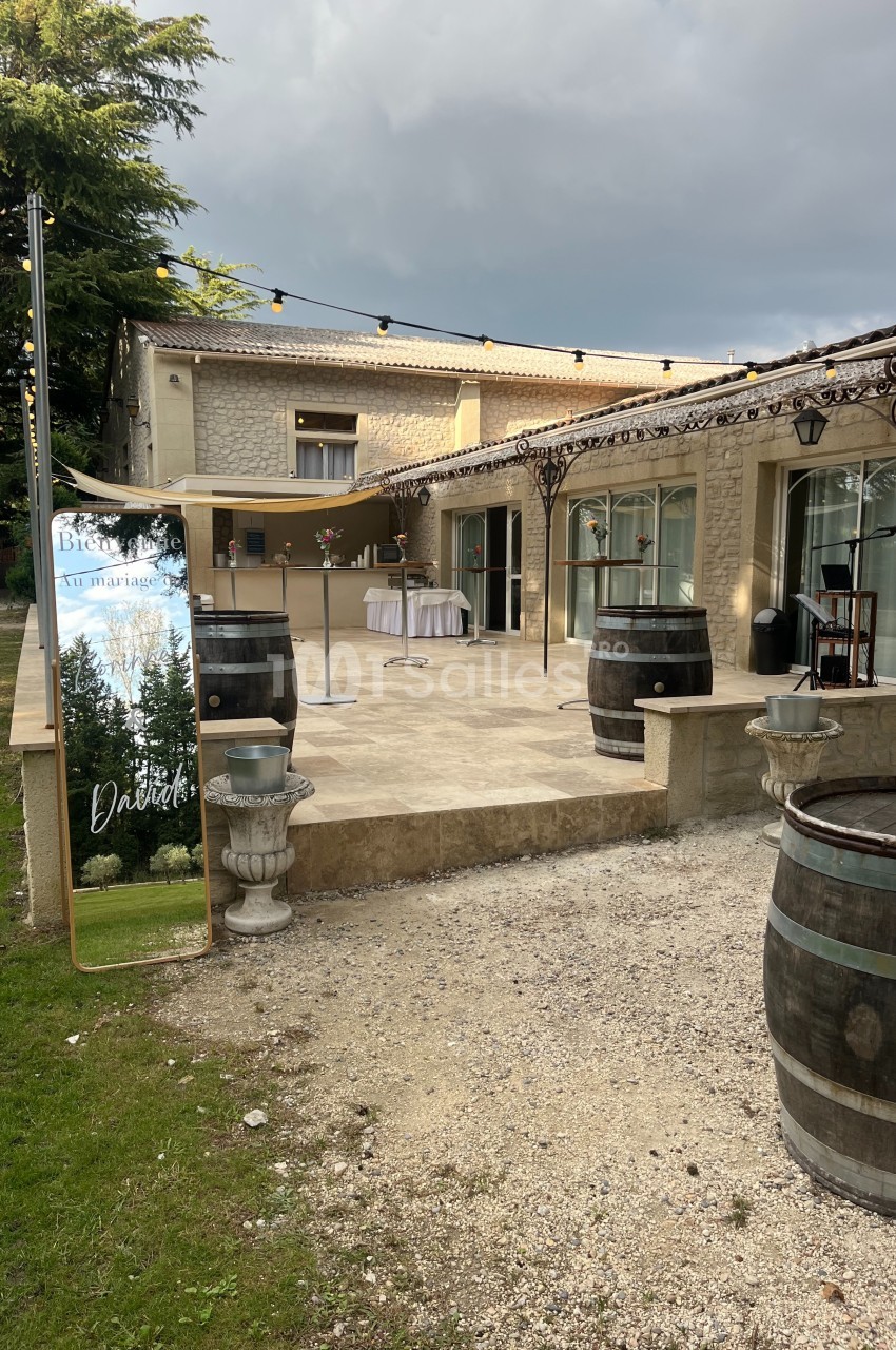 Terrasse d'un bâtiment en pierre avec barils en bois, guirlandes lumineuses et panneau décoratif pour un événement.