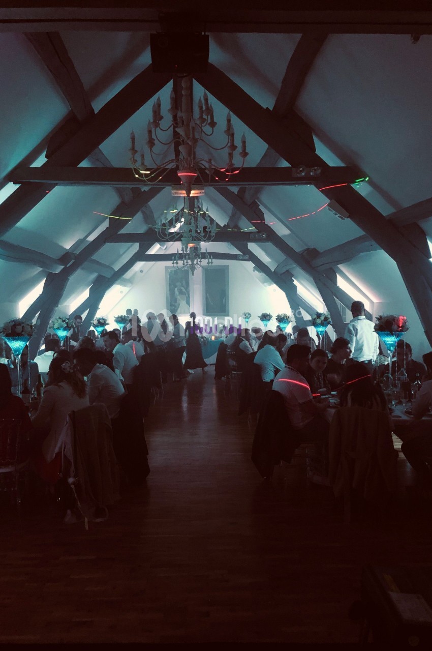 Salle de réception avec des invités assis à des tables décorées, éclairage tamisé et ambiance festive.