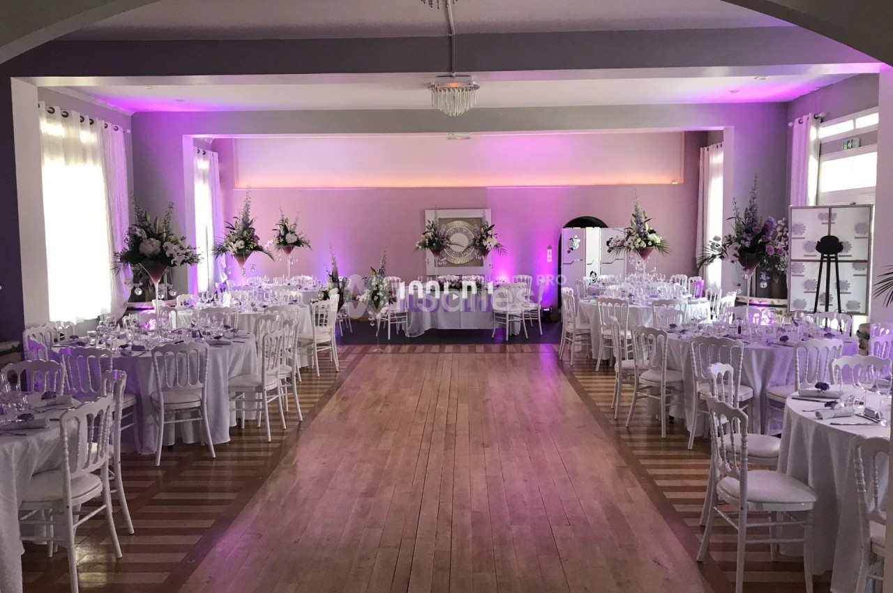Salle de réception décorée avec des tables rondes, des chaises blanches et un éclairage rose.