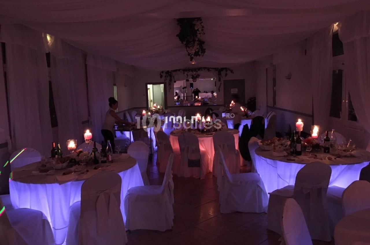 Salle de réception décorée avec des tables rondes éclairées en bleu, nappes blanches et bougies allumées.