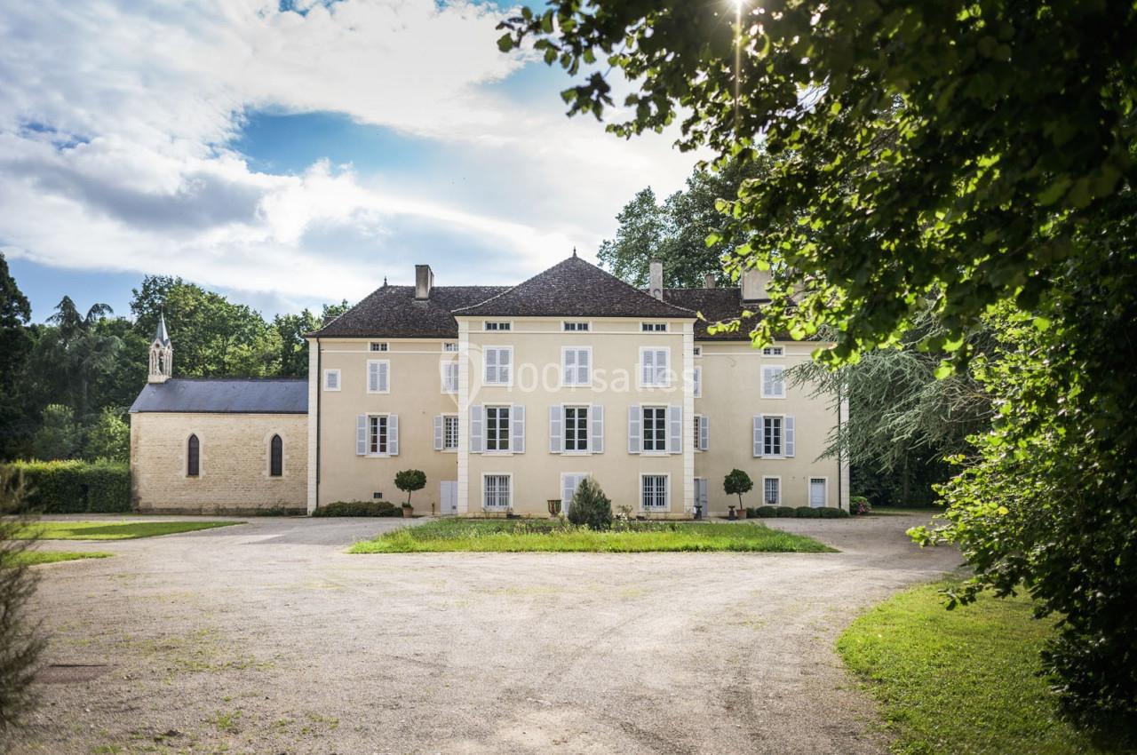 Façade d'un manoir jaune clair avec une chapelle attenante, entouré de verdure sous un ciel partiellement nuageux.