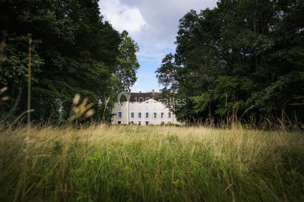 Manoir blanc entouré d'arbres, vu à travers une prairie verdoyante sous un ciel partiellement nuageux.