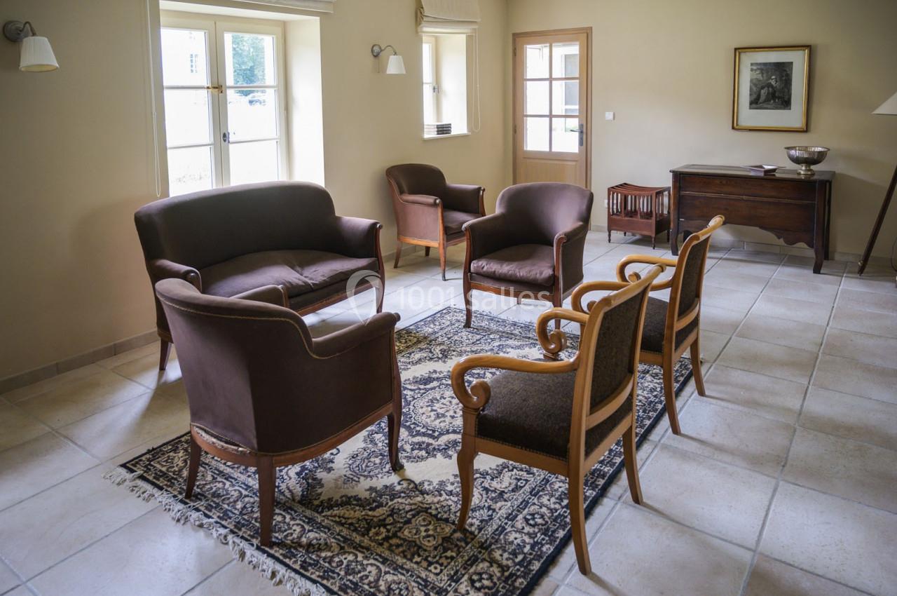 Salon lumineux avec mobilier en bois et tissu brun, tapis à motifs, carrelage clair et fenêtres donnant sur l'extérieur.