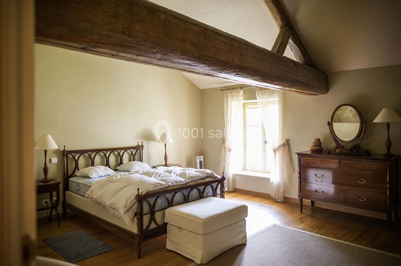 Chambre lumineuse avec lit double, poutre apparente, commode en bois, fauteuil et fenêtre avec rideaux clairs.