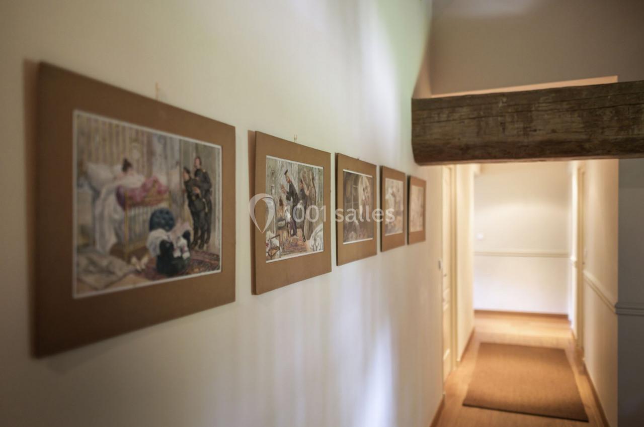 Couloir éclairé avec des cadres illustrés accrochés au mur, poutre en bois apparente au plafond.