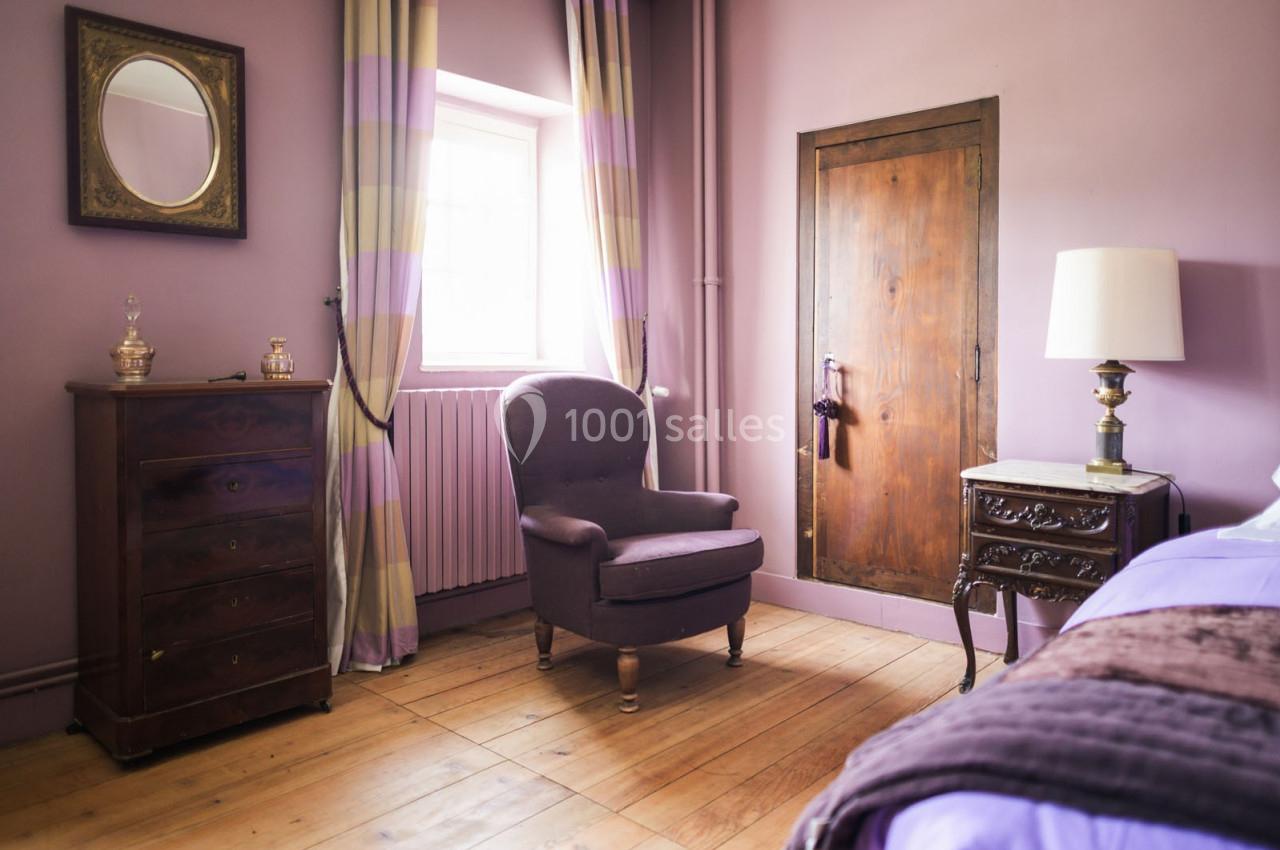 Chambre avec murs mauves, fauteuil, commode, lampe sur table et fenêtre laissant entrer la lumière naturelle.