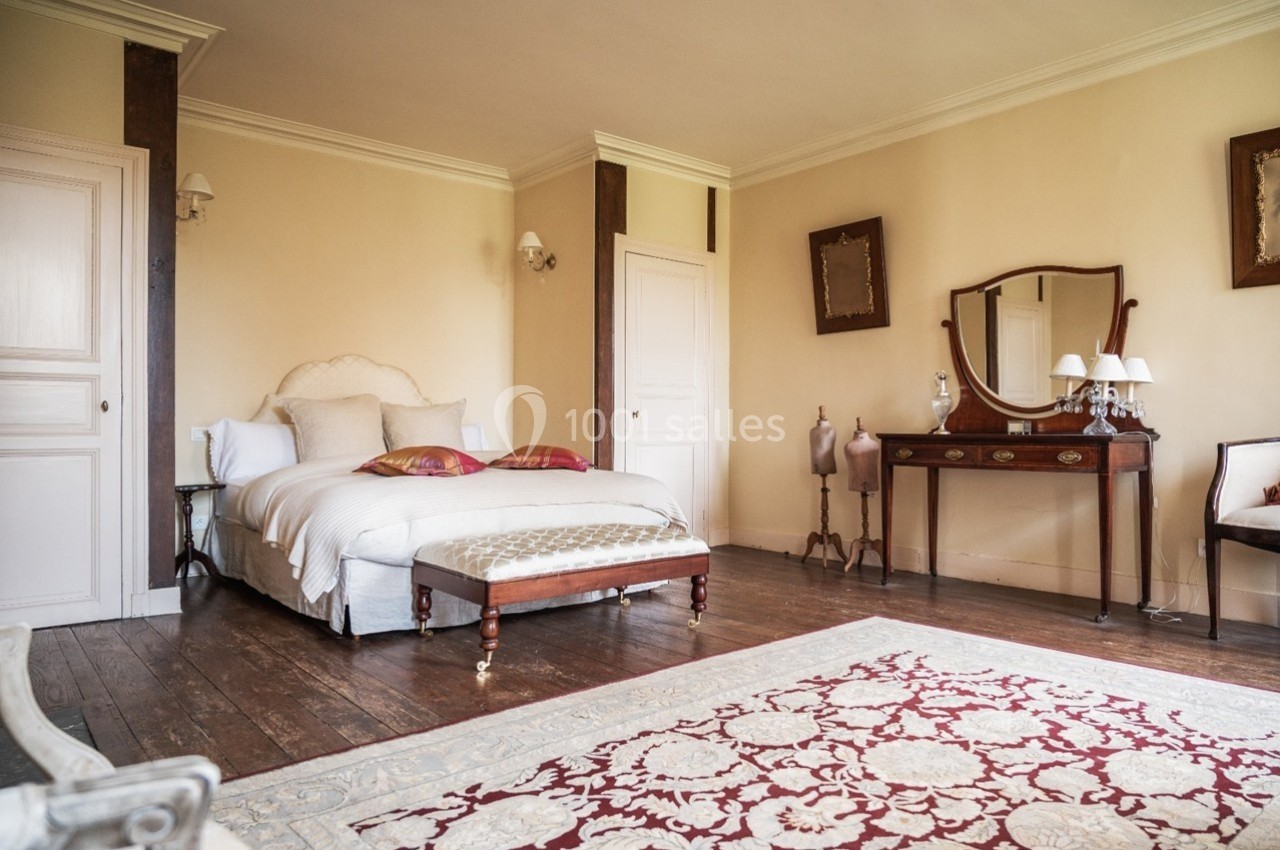 Chambre lumineuse avec lit double, mobilier ancien en bois, tapis décoratif et murs beige clair.