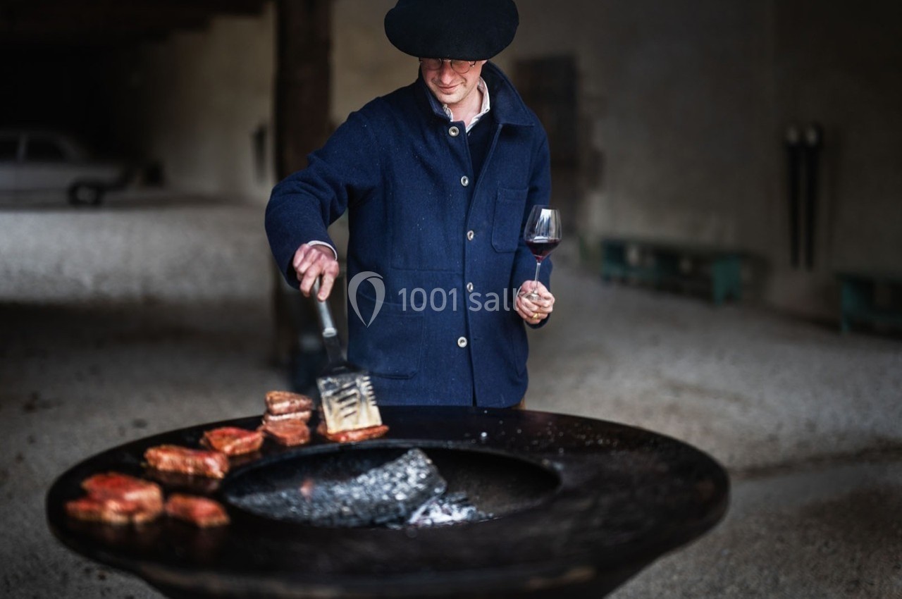 Un homme en veste bleue et chapeau cuisine des morceaux de viande sur un brasero, un verre de vin à la main.