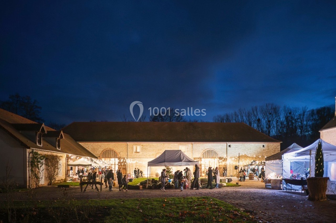 Cour d'une ferme illuminée en soirée, avec des stands, des lumières et des visiteurs rassemblés.