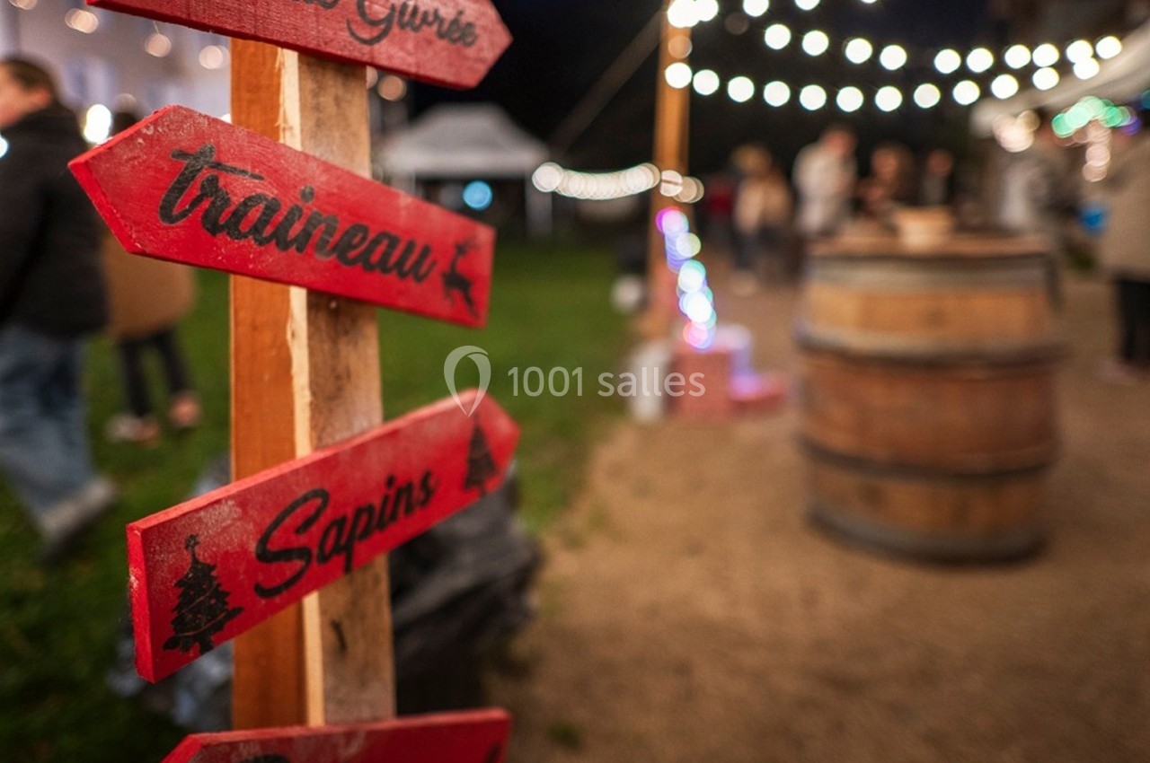 Panneaux rouges en bois indiquant ’traîneau’ et ’sapins’ dans un marché nocturne éclairé par des guirlandes lumineuses.