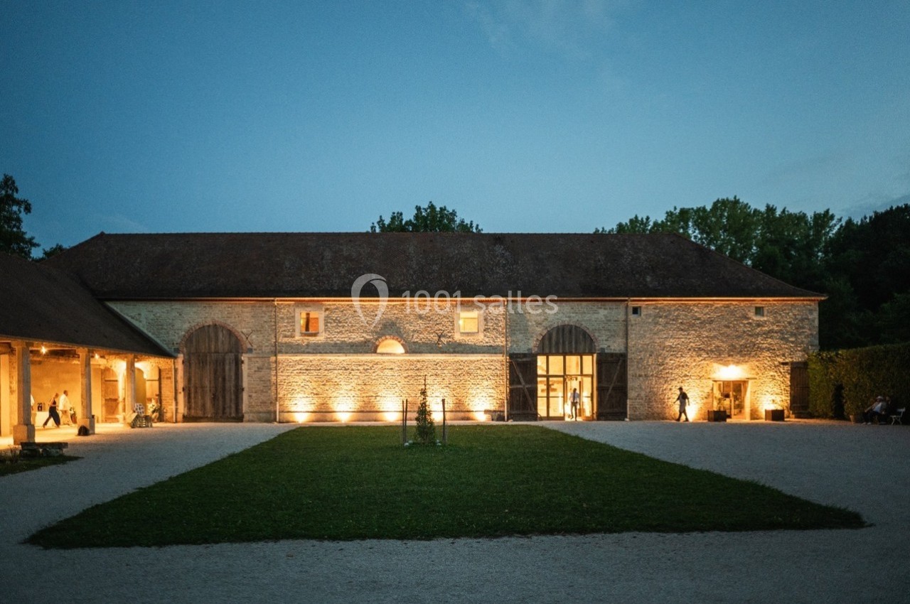 Bâtiment en pierre éclairé de nuit, entouré d'une cour avec pelouse et allées gravillonnées.