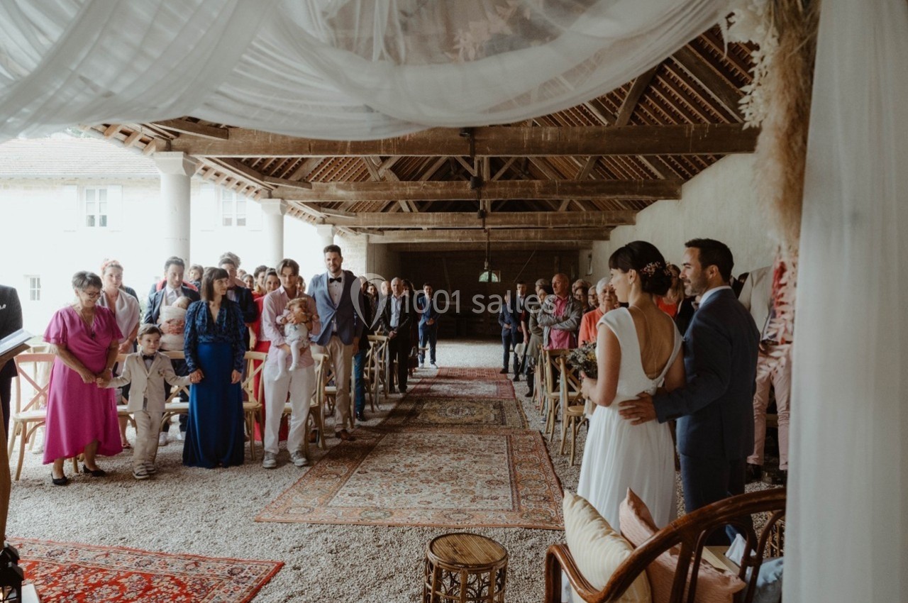 Un couple se tient devant des invités assis dans une salle de cérémonie décorée avec des tapis et des voilages.