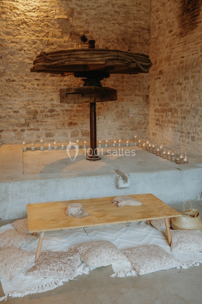Ancienne presse en pierre dans une pièce en pierre, entourée de bougies, avec une table basse et des coussins au premier…