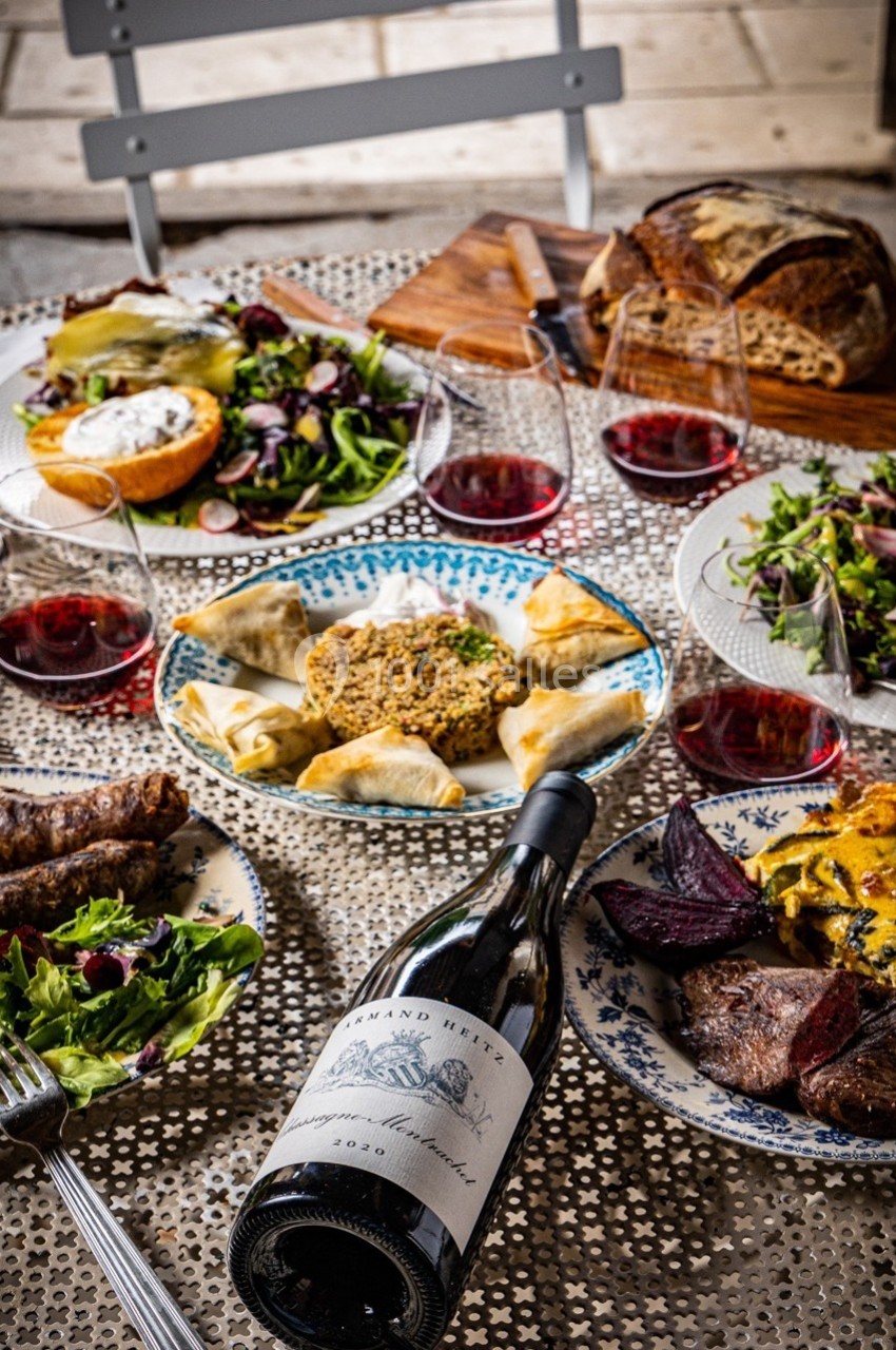 Table dressée avec des plats variés, salades, pain, vin rouge et mets cuisinés, sur une nappe ajourée.