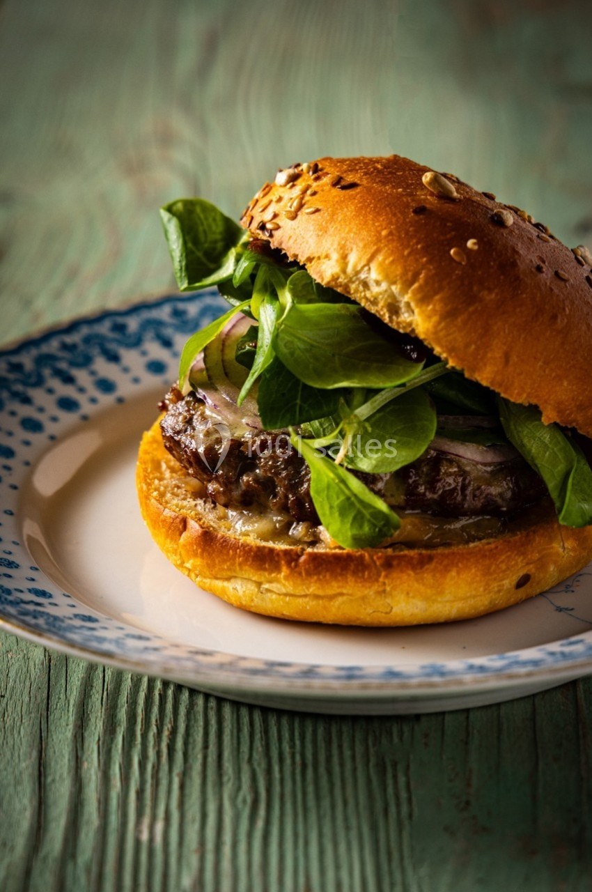 Burger garni de mâche, oignons rouges et fromage fondu, servi dans un pain aux graines sur une assiette blanche et bleue.