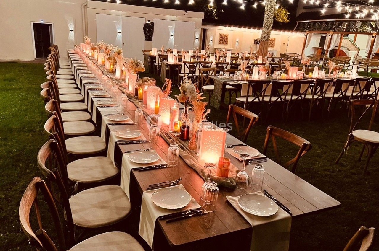 Grande table en bois décorée de bougies et fleurs, alignée sous des guirlandes lumineuses dans un jardin en soirée.