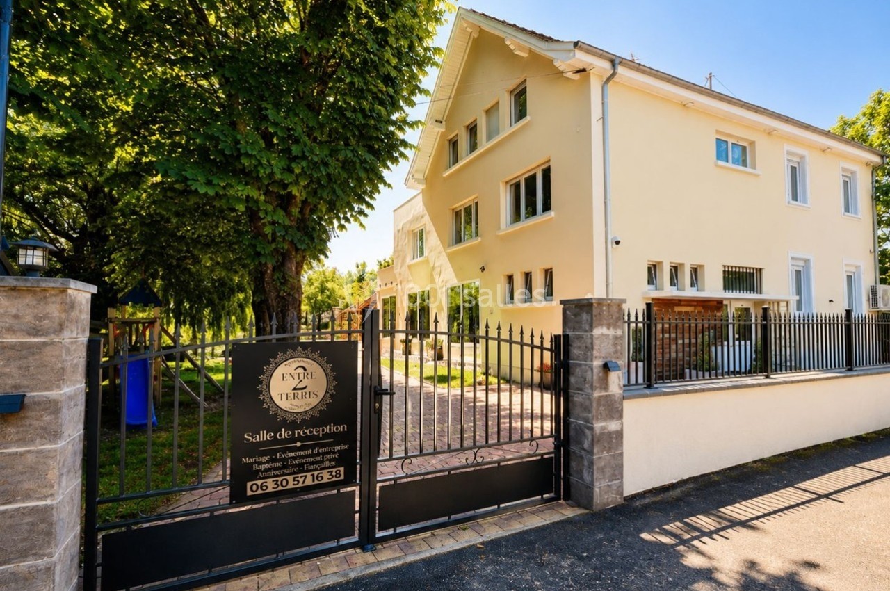 Grande maison beige entourée d'arbres, avec une entrée clôturée et un panneau indiquant une salle de réception.