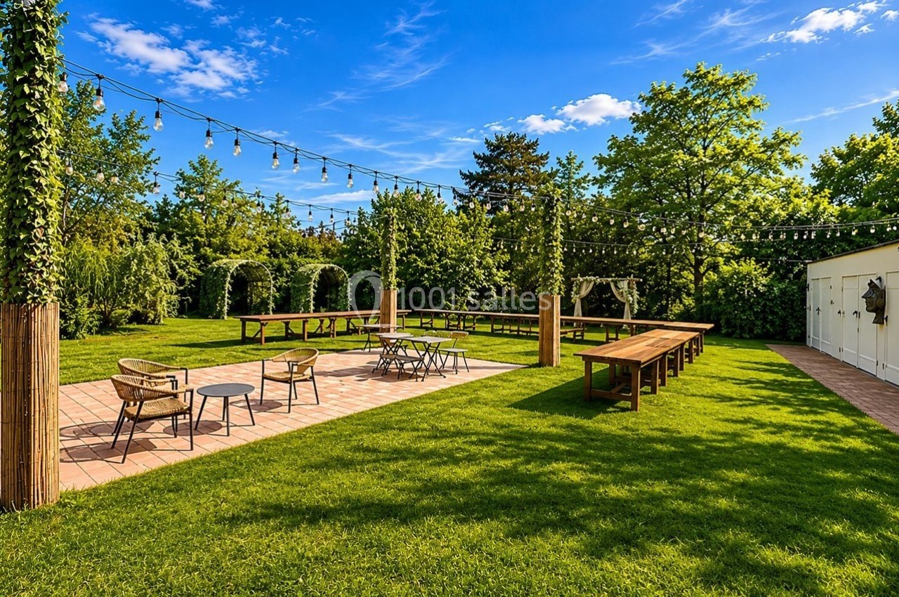 Jardin aménagé avec tables en bois, chaises, guirlandes lumineuses suspendues et allée pavée entourée de verdure.