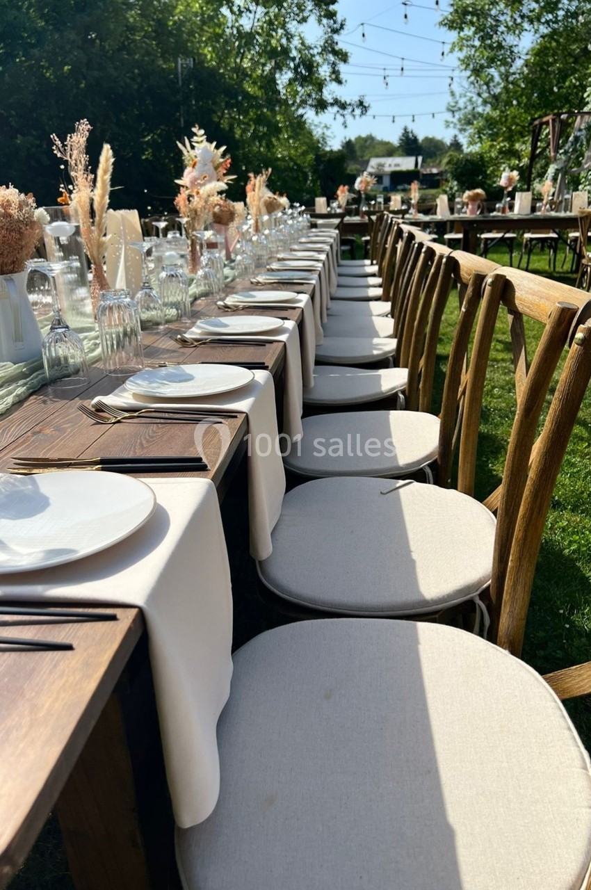 Table en bois dressée en extérieur avec nappes blanches, assiettes, verres et décorations florales sous des guirlandes…