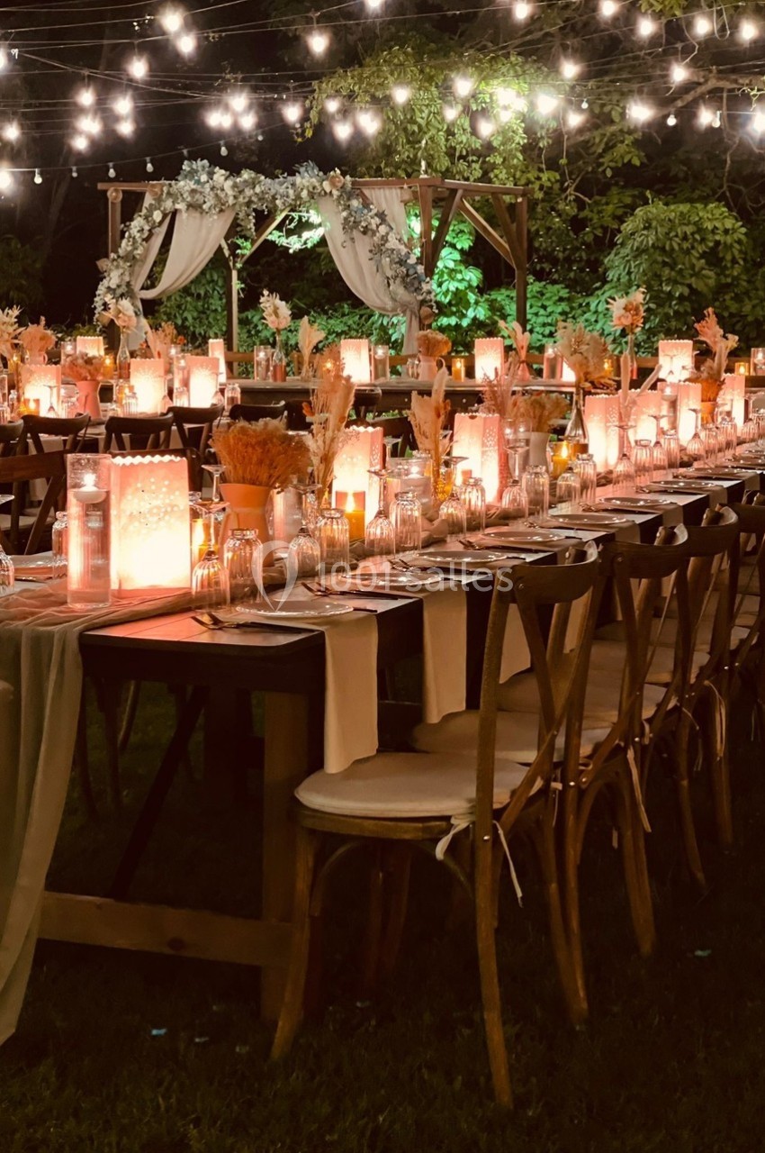Tables décorées pour un dîner en extérieur, éclairées par des guirlandes lumineuses et ornées de fleurs et bougies.