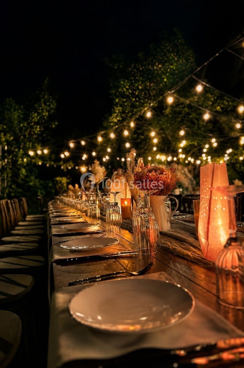 Grande table en bois décorée pour un dîner en extérieur, éclairée par des guirlandes lumineuses et des bougies.