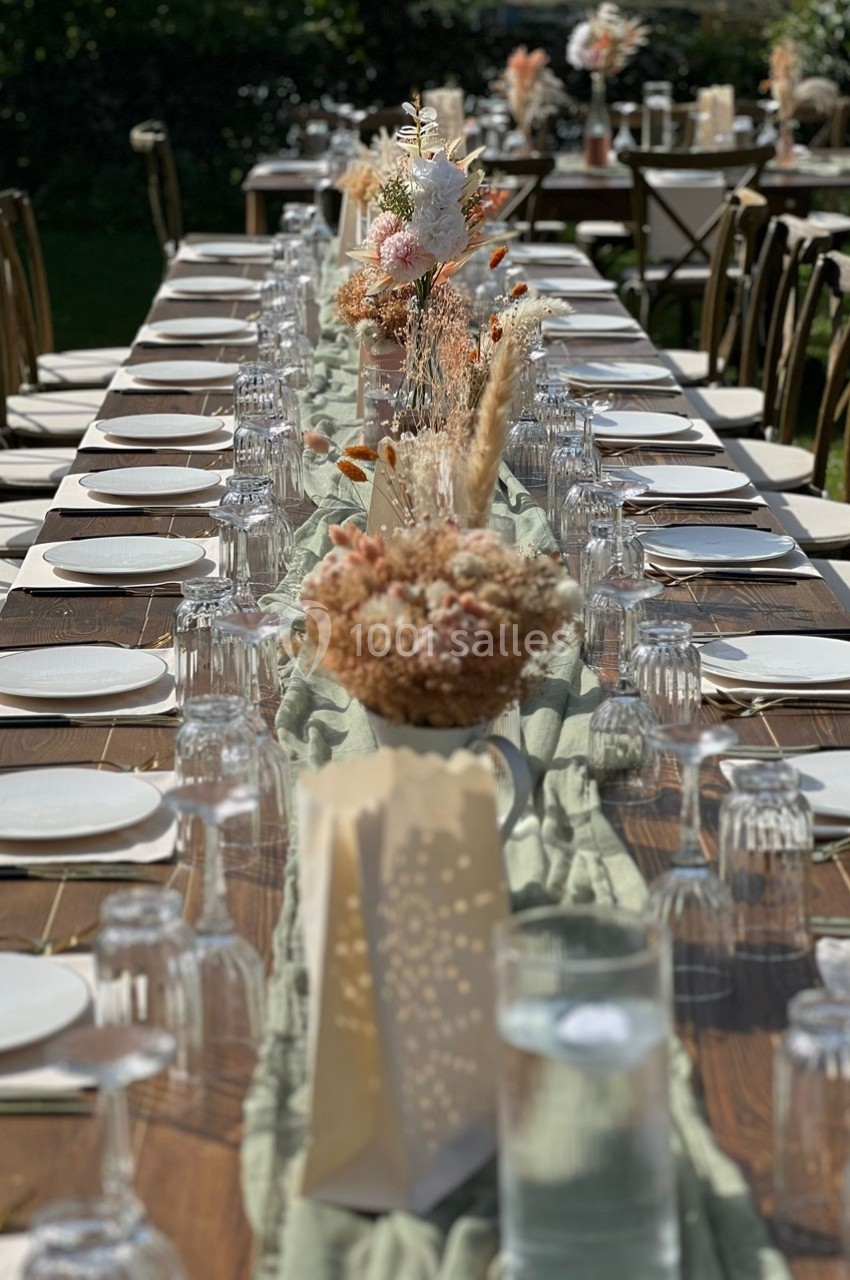 Grande table en bois dressée pour un repas en extérieur, décorée de fleurs séchées et de vaisselle élégante.