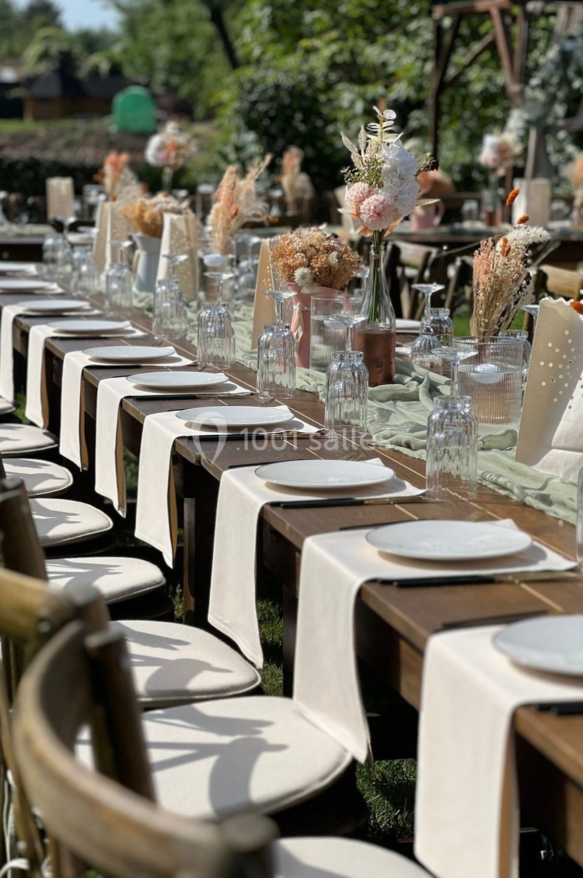 Grande table extérieure décorée pour un événement, avec nappes blanches, assiettes, verres et bouquets de fleurs séchées.
