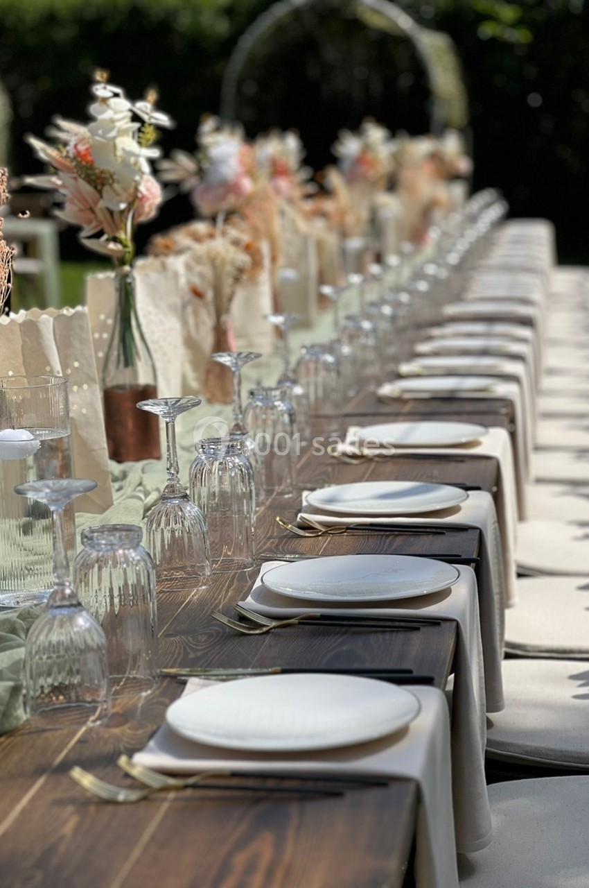Table en bois dressée pour un événement, avec assiettes blanches, verres et décoration florale dans un cadre extérieur.
