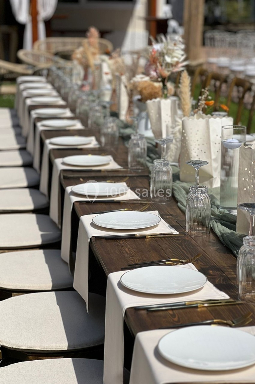 Table en bois dressée pour un repas, avec assiettes blanches, verres et décorations florales dans un cadre extérieur.