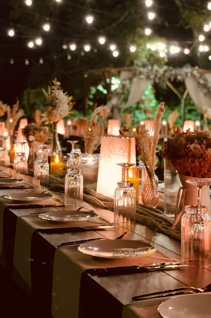 Table décorée pour un dîner en extérieur, éclairée par des guirlandes lumineuses et ornée de fleurs et bougies.