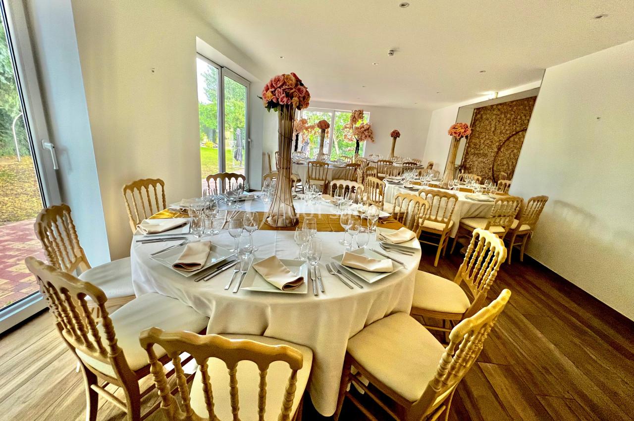 Salle de réception lumineuse avec tables rondes dressées, chaises en bois doré et décorations florales.