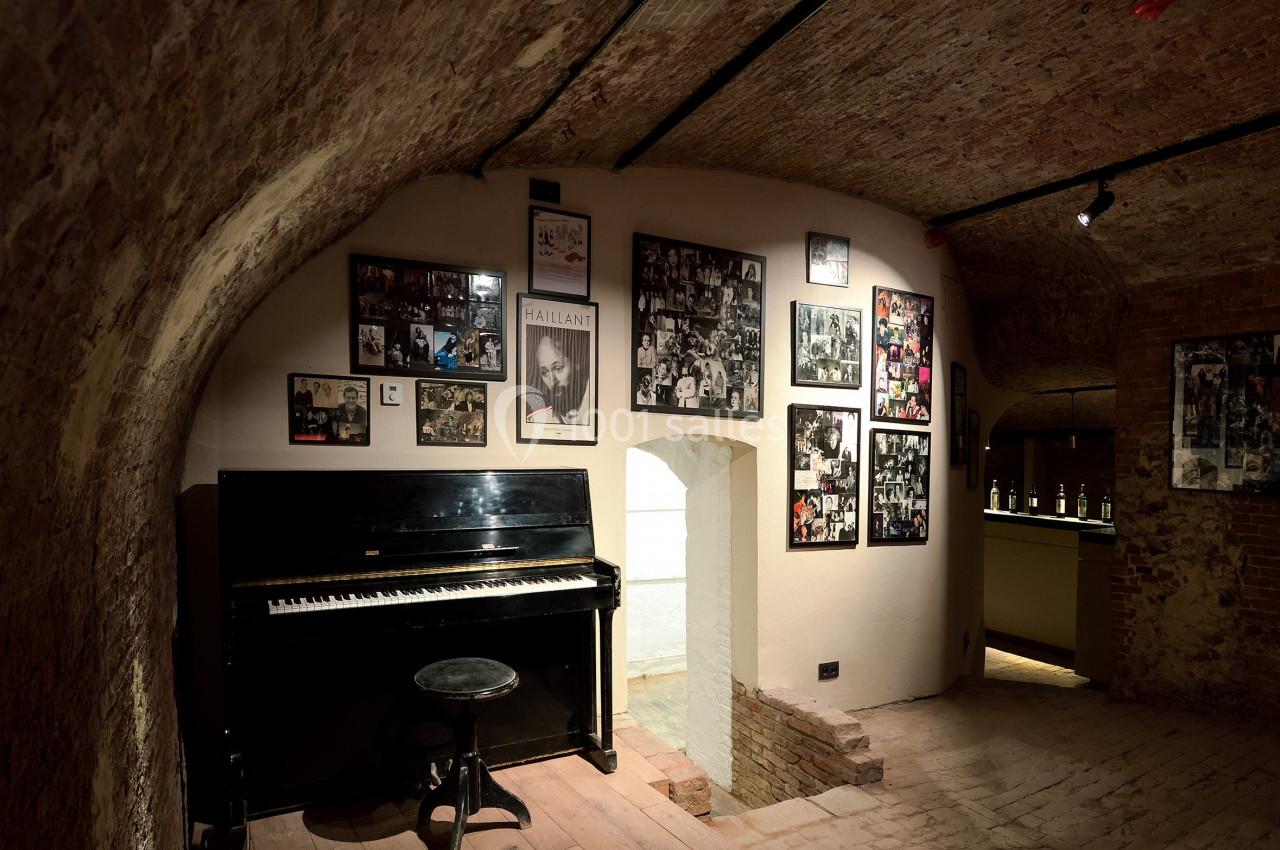 Piano noir avec tabouret dans une pièce voûtée en briques, décorée de photos encadrées sur les murs.